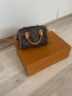 Louis Vuitton Monogram Speedy Nano - Superfin Louis Vuitton Speedy Nano i klassiskt brunt monogramcanvas med ljusbruna läderdetaljer och guldfärgade metalldelar. Väskan har både handtag och en lång axelrem, perfekt för att bära crossbody. Ikonisk design med dragkedja upptill och snygg form.