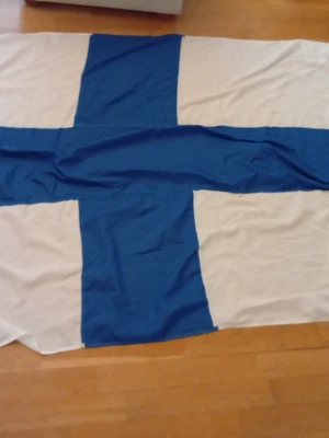 Finlands flagga i tyg - Stor tygflagga med blått kors på vit bakgrund, klassisk design för Finlands flagga. Perfekt för att visa stöd vid sportevenemang eller som dekoration. Rektangulär form och tillverkad i lätt tygmaterial.