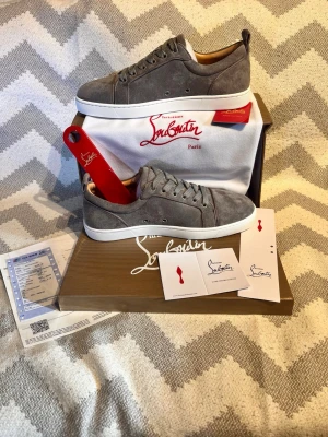 Gråa Louboutin sneakers i mocka - Snygga grå sneakers från Christian Louboutin i mjuk mocka med klassisk röd sula och vita kanter. Skorna har lågt skaft, rund tå och matchande grå skosnören. Perfekta för dig som vill sticka ut med exklusiv stil. (Allt på bilderna medföljer)