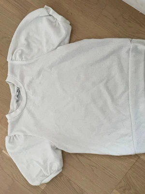 Vit topp med puffärm från 157 - Vit topp från 157 med korta puffärmar och rund halsringning. Tillverkad i mjukt bomullsmaterial som känns skönt mot huden. Enkel och stilren design som passar till många olika outfits.