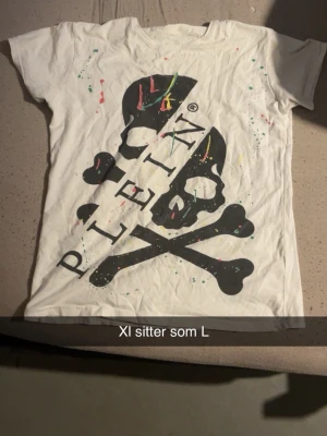 Vit Plein t-shirt med döskalletryck Storlek Xl men sitter mer som en L - Säljer en vit t-shirt från Plein med stort svart döskalle- och korslagda ben-tryck framtill samt färgstänk i rosa, gul och blå. T-shirten har korta ärmar och normal passform. Perfekt för dig som gillar statement-plagg och streetwear vibes.