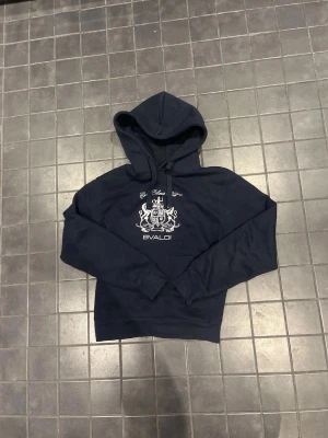 Mörkblå hoodie från Bvaldi - Mörkblå hoodie från Bvaldi med vit tryckt logga och emblem på bröstet. Klassisk huva med dragsko och långa ärmar. Perfekt för en avslappnad och cool stil. Materialet känns mjukt och bekvämt mot huden.