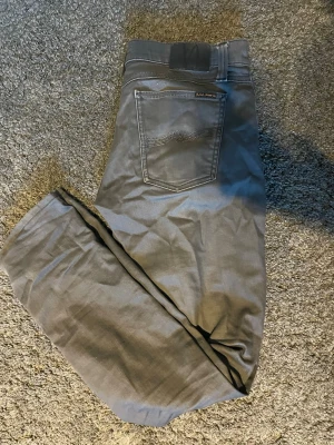 Gråa jeans från nudie jeans - Säljer ett par nudie jeans i färgen moog grey i modellen tight long john i storlek W33 L32  med klassisk femficksdesign och raka ben. Jeansen har en diskret logotyp på bakfickan och är tillverkade i ett mjukt bomullsmaterial med lite stretch för extra komfort.