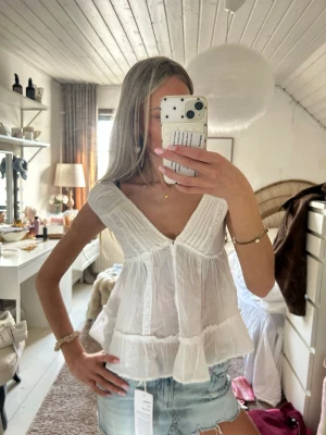 Vit volangblus med v-ringning - Söt vit blus med djup v-ringning och breda axelband. Blusen har volangdetaljer nedtill och ett luftigt, lätt material som passar perfekt till sommaren. Stilren och enkel att matcha till jeans eller kjol.