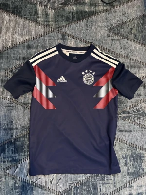 Storlek 150cm Adidas Bayern München träningströja 2021-22 - Mörkblå träningströja från Adidas med Bayern München-logga och vita samt röda detaljer på bröstet. Tröjan har korta ärmar, rund hals och klassiska vita Adidas-ränder på axlarna. Tillverkad i ett lätt och funktionellt material som passar perfekt till träning. 