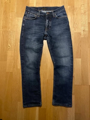 Mörkblå jeans från Nudie Jeans - Snygga mörkblå jeans från Nudie Jeans i modellen ”Dude Dan” med klassisk femficksdesign och orangea sömmar på bakfickorna. Jeansen har en rak passform och är tillverkade i denim av bomull. Perfekta för en avslappnad och trendig stil. Dock en liten skada på höger bakficka, se bild 5. Fråga gärna om du undrar något! Mått: Längd 96 cm, Midja 39 cm