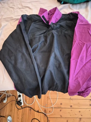 Svart och lila retro vindjacka - Cool vindjacka i svart och lila med hög krage och dragkedja. Oversized passform och klassisk retrostil, perfekt för att sticka ut. Jackan har elastisk nederkant och är tillverkad i ett lätt, vindtätt material.