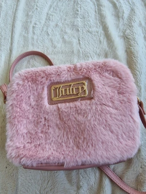 Rosa fluffig axelväska Juicy Couture  - Superfin rosa axelväska från Juicy med mjuk fuskpäls och guldfärgad logga framtill. Väskan har justerbar axelrem och flera fack inuti för enkel organisering. Perfekt för dig som gillar en söt och trendig look.