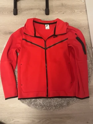 Röd Nike hoodie med dragkedja (äkta) - Snygg röd hoodie från Nike med hel dragkedja framtill och svarta detaljer. Hoodien har en huva och praktiska fickor på sidorna, samt Nike-logga på framsidan. Tillverkad i mjukt material som känns skönt mot huden. Perfekt för en sportig och avslappnad stil.