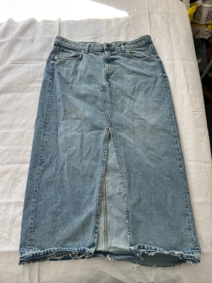 Lång jeanskjol med slits framtill - Trendig lång jeanskjol i ljusblå denim med rå, fransig fåll och slits framtill. Klassisk femficksmodell med bälteshällor och dragkedja. Perfekt för dig som gillar en avslappnad och cool stil.