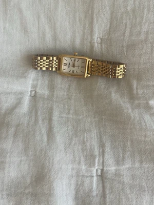 Guldfärgad rektangulär-klocka - Stilren guldfärgad klocka  med rektangulärt vitt ur och romerska siffror. Länken är i metall med klassisk design och klockan har en elegant look som passar till många outfits.