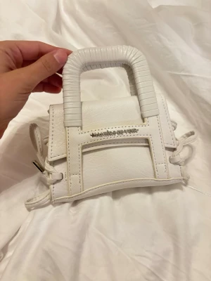 Vit miniväska från Maniere De Voir - Superfin vit minihandväska från Maniere De Voir i stilren design. Väskan har ett fyrkantigt handtag, detaljerade sömmar och snörning på sidorna. Tillverkad i slätt läder med logga framtill. Perfekt för dig som gillar trendiga accessoarer.