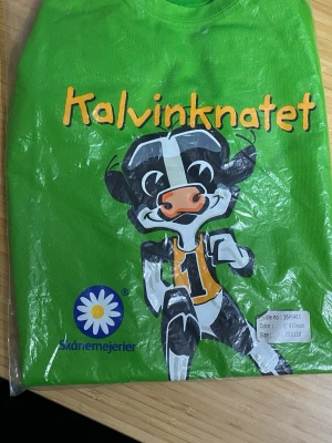 Grön Kalvinknatet t-shirt  - Grön t-shirt med tryck av Kalvinknatet och en tecknad ko på framsidan. T-shirten har rund hals och korta ärmar. Perfekt för dig som gillar färgglada och lekfulla plagg. Officiell merch från Skånemejerier.