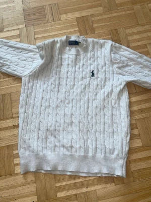 Vit kabelstickad tröja Polo Ralph Lauren - Kabelstickad vit tröja från Polo Ralph Lauren med rund halsringning och klassisk broderad logga på bröstet. Tröjan har ribbade muddar vid ärmslut och nederkant. Perfekt för en stilren och tidlös look.
