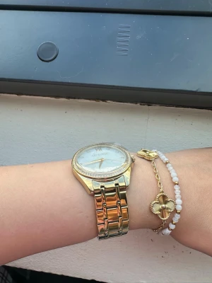 Guldig klocka från Ivy N Rox - Stilren klocka från Ivy N Rox i guldigt metallarmband och boett. Urtavlan är vit med romerska siffror och små stenar som index. Runt boetten sitter glittriga stenar som ger extra lyxig känsla. Helt ny och oanvänd, plasten kvar 