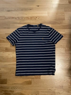 Randig t-shirt från Polo Ralph Lauren - Mörkblå t-shirt med vita horisontella ränder från Polo Ralph Lauren. Klassisk passform med korta ärmar och rund hals. Liten broderad logga på bröstet. Tillverkad i mjuk bomull som känns skön mot huden. Storleken är L men skulle säga att den även passsr M! 