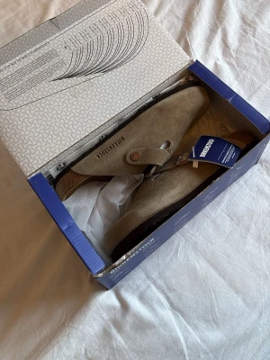 Birkenstock Boston taupe tofflor - Säljer ett par Birkenstock Boston tofflor i färgen taupe. De är tillverkade i mjukt mockaläder med justerbart spänne och klassisk korksula för skön komfort. Perfekta för dig som gillar stilrena och tidlösa skor. Levereras i originalkartong. De är aldrig använda allting som nytt😁