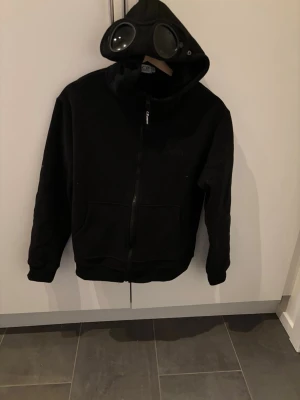 Cp company hoodie - Cool svart hoodie från C.P. Company med hel dragkedja och ikoniska goggles i huvan. Hoodien har två stora fickor framtill och broderad logga på bröstet. Perfekt för dig som vill sticka ut med en unik streetwear-look.