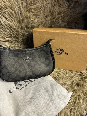 Svart axelväska  - Snygg svart axelväska med klassiskt monogrammönster och silverfärgade detaljer. Väskan har en smidig dragkedja upptill och är tillverkad i tåligt konstläder. Perfekt storlek för det viktigaste och en ikonisk look.