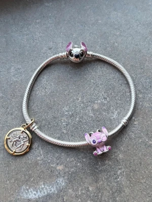 Silverfärgat armband med berlocker - Snyggt armband i silverfärg med två färgglada berlocker som föreställer figurer i rosa och lila samt ett runt hänge i guld- och silverton med detaljerat mönster. Perfekt accessoar för dig som gillar unika smycken och vill sticka ut. Går att förhandla pris.