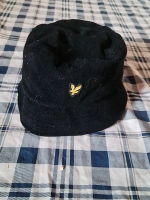 Svart bucket hat från Lyle & Scott - Svart bucket hat från Lyle & Scott med klassisk gul logga framtill. Hatten är i mjukt tyg och har en avslappnad form som passar perfekt till streetwear eller casual stil.