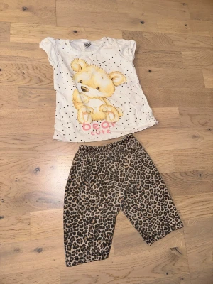 Sött set med björntopp och leopardbyxor - Sött set med björntopp och leopardbyxorTvådelat set med en vit t-shirt med svartprickigt mönster och en stor söt björn på framsidan samt texten 'bear cute'. Tillhörande byxor i leopardmönster med resår i midjan. Perfekt för dig som gillar gulliga och trendiga prints.