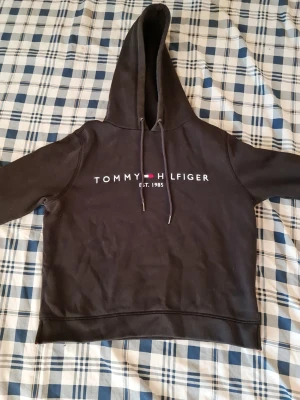 Svart hoodie från Tommy Hilfiger - Snygg svart hoodie från Tommy Hilfiger med klassisk logga och texten 'EST. 1985' på bröstet. Huva med dragsko och ribbade muddar. Perfekt för en chill och stilren look.