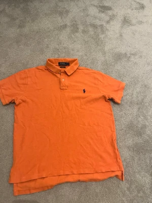 Orange pikétröja från Polo Ralph Lauren - Säljer en orange pikétröja från Polo Ralph Lauren i storlek L. Tröjan har klassisk krage, korta ärmar och knappar vid halsen. Ikoniska loggan broderad på bröstet. Tillverkad i bomull och har en normal passform.
