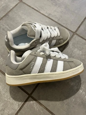 Adidas grå Gazelle sneakers - Snygga Adidas Gazelle sneakers i grå mocka med klassiska vita ränder på sidorna och vit sula. Skorna har lågt skaft, breda vita skosnören och gummisula i ljusbrun färg. Perfekta för dig som gillar en clean och sportig stil.