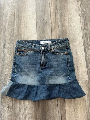 Jeanskjol med volang  - Snygg blå jeanskjol med volang nertill och klassiska fickor fram och bak. Kjolen har bälteshällor, knapp och dragkedja framtill. Perfekt för dig som gillar en trendig och avslappnad stil.