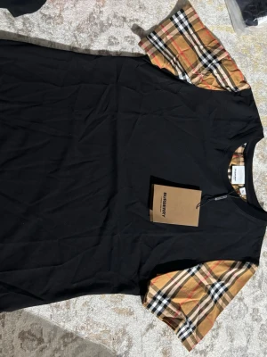 Burberry t-shirt - Svart t-shirt från Burberry med klassiska rutiga korta ärmar i beige, svart, vitt och rött. T-shirten har rund halsringning och en stilren design som sticker ut med de ikoniska ärmarna. Perfekt för dig som vill ha en trendig och exklusiv look.