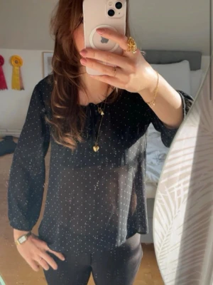 Svart prickig blus med knytband - Snygg svart blus med vita småprickar och transparent material. Blusen har långa ärmar och ett knytband vid halsen för en extra detalj. Perfekt att styla med jeans eller kjol för en trendig look.