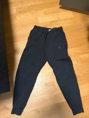 Svarta mjukisbyxor från Nike (tröja finns) - Går att köpa med tröja Säljer ett par svarta mjukisbyxor från Nike med dragsko i midjan och ribbade muddar vid bensluten. Byxorna har en avslappnad passform och diskret Nike-logga på benet. Perfekta för chill dagar eller träning.