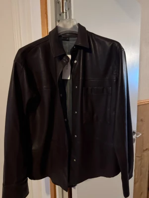 Mörkbrun skinnjacka med skjortkrage - Stilren mörkbrun skinnjacka med skjortkrage och tryckknappar framtill. Jackan har en bröstficka och raka ärmar, perfekt för en trendig och avslappnad look. Materialet är mjukt skinn som ger en lyxig känsla. Nypris är 500kr köpte min för 250 och säljer den för 150. Aldrig använd!