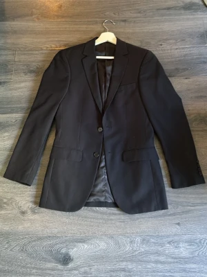 Svart klassisk kavaj från dressman - Stilren svart kavaj med två knappar framtill och klassisk passform. Kavajen har två fickor med lock, bröstficka och helfodrad insida. Perfekt för en dressad look och passar till både skjorta och t-shirt.