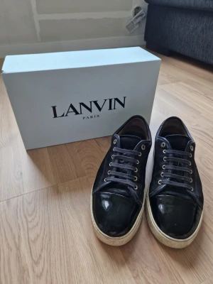 Svarta Lanvin sneakers med lackdetaljer - Snygga svarta sneakers från Lanvin med klassisk låg profil. Skorna har en unik kombination av mocka och glansiga lackdetaljer på tån, samt vita sulor och metallfärgade öljetter. Perfekta för dig som vill ha en stilren och exklusiv look.