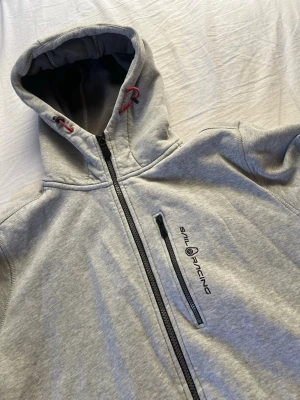 Grå hoodie från Sail Racing - Grå hoodie från Sail Racing med dragkedja framtill och svart logga på bröstet. Hoodien har en bröstficka med dragkedja, justerbar huva med röda detaljer och mjukt material. 