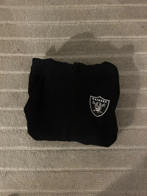 Svart Raiders hoodie med tryck - Snygg svart hoodie med Raiders-logga på bröstet och stort RAIDER-tryck på ryggen. Klassisk huva och långärmad modell, perfekt för dig som gillar sportig streetstyle. Mjuk bomullskänsla och enkel att matcha med jeans eller joggers.