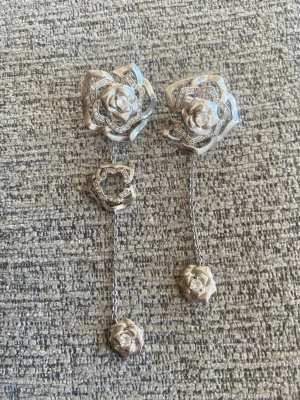 Silvriga örhängen med rosdesign - Unika örhängen i silverfärg med rosformade detaljer och gnistrande stenar. Örhängena har en cool kedja som binder ihop två rosor på varje örhänge, vilket ger en trendig och elegant look. Perfekta för dig som gillar smycken med det lilla extra. Från ioaku