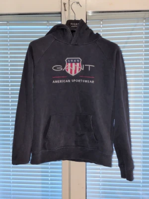 Svart hoodie från Gant med tryck - Snygg svart hoodie från Gant med stort tryck på bröstet och texten 'American Sportswear'. Klassisk känguruficka framtill och huva. Mjuk bomullskänsla och avslappnad passform, perfekt för en casual look.