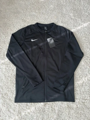 Svart träningsjacka från Nike - Svart träningsjacka från Nike med hel dragkedja och två sidofickor med dragkedja. Jackan har en vit Nike-logga på bröstet och är tillverkad i ett slätt, sportigt material som passar perfekt till träning eller uppvärmning.