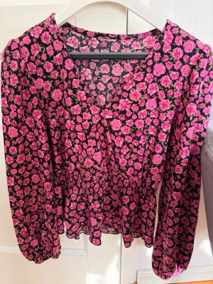 Blommig svart blus med rosa rosor - Supersöt blus med svart botten och tätt mönster av rosa rosor. Blusen har v-ringning, markerad midja och långa ärmar med resår vid ärmslut. Tunt och luftigt material som ger en flowy känsla. Perfekt för dig som gillar färg och mönster!