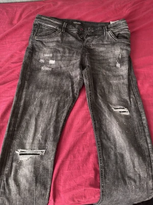 Grå slitna jeans med hål - Säljer ett par grå jeans från Jack & Jones med slitningar och hål på benen. Jeansen har fem fickor, knappgylf och en cool tvättad look. Perfekta för dig som gillar en avslappnad och trendig stil.