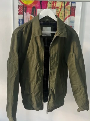 Olivgrön jacka från Pull&Bear - Snygg olivgrön jacka från Pull&Bear med klassisk krage och dragkedja. Jackan har två fickor framtill och rutigt innerfoder i mörkblått, rött och vitt. Perfekt för dig som gillar en stilren och enkel look.