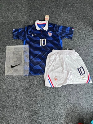 Mbappe Frankrike fotbollströja och shorts - Säljer ett matchställ från Frankrikes landslag med blå fotbollströja och vita shorts. Tröjan har nummer 10, guldigt Nike-märke och FFF-logga med tupp. Shortsen är vita med blå och röda detaljer samt nummer 10. Materialet är lätt och andas, perfekt för fotboll. L/M storlek