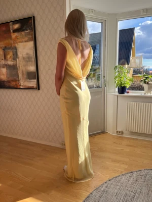 Ljusgul långklänning med öppen rygg - Elegant ljusgul långklänning med vacker öppen rygg och draperad axel. Klänningen har en mjuk, flytande siluett och är tillverkad i ett tunt, lätt material som ger en lyxig känsla. Perfekt för dig som vill sticka ut med en stilren och sofistikerad look.