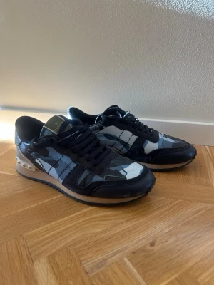 Valentino Rockrunners blåa svarta - Snygga sneakers från Valentino med svart och blått camo-mönster, svarta mockadetaljer och coola nitar på hälen. Skorna har klassisk låg profil och vita samt svarta detaljer på sulan. Perfekta för dig som vill sticka ut med stil.