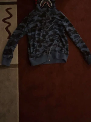 Blå Bape hoodie med full zip upp - Säljer en blå camouflagemönstrad hoodie från BAPE med dragkedja och huva. Huvan har ett hajmotiv med tänder och ögon, vilket ger en unik streetwear-look. Hoodien har ribbade muddar och är perfekt för dig som gillar statement-plagg.