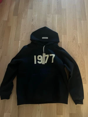 Svart Essentials 1977 hoodie - Snygg svart hoodie från Essentials med stor vit '1977'-print på bröstet och klassisk huva. Hoodien har en relaxed fit och är tillverkad i mjukt bomullsmaterial. Perfekt för dig som gillar streetwear och vill ha en clean look.