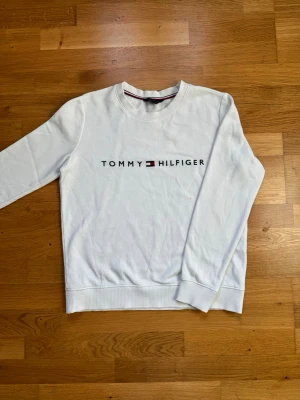 Vit sweatshirt från Tommy Hilfiger - Vit sweatshirt med rund hals och lång ärm från Tommy Hilfiger. Tröjan har en broderad logotyp i svart, rött och blått framtill. Klassisk och stilren design med ribbade muddar vid ärmslut och nederkant. Passar även storlek S